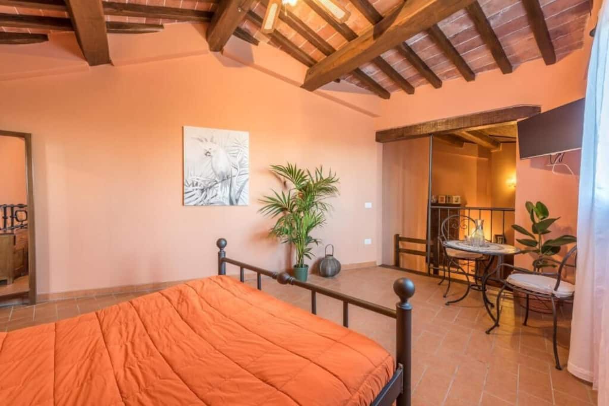 A bedroom with a bed, a potted plant, a painting, and a balcony with a table and chairs.