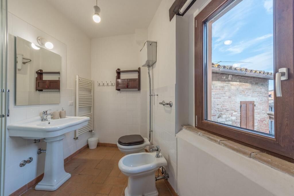 Une salle de bain équipée d’un lavabo, de toilettes, d’un bidet et d’une fenêtre donnant sur un immeuble en briques.