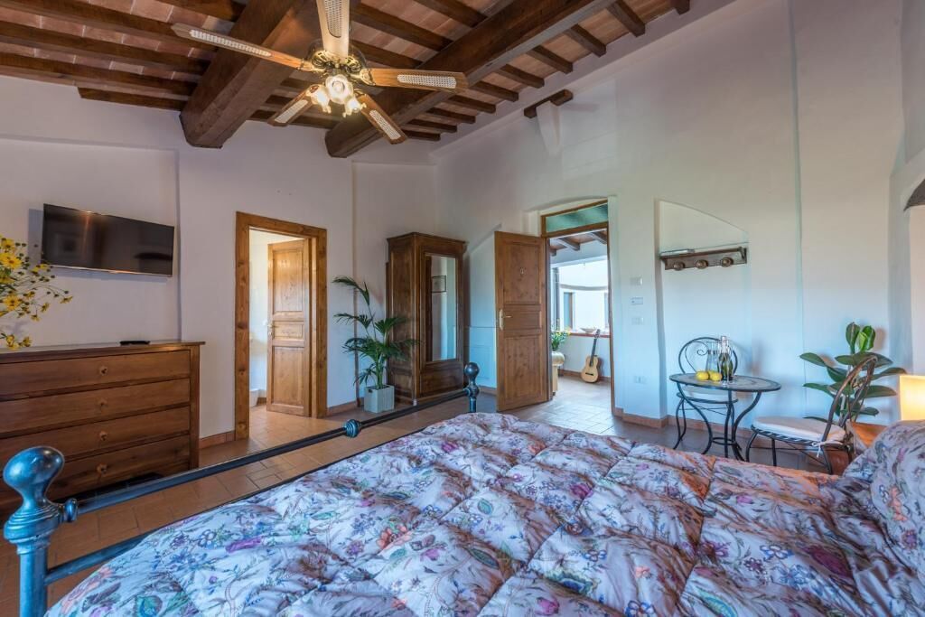 A bedroom with a bed, wooden ceiling beams, a ceiling fan, a TV, and a door leading to another room.