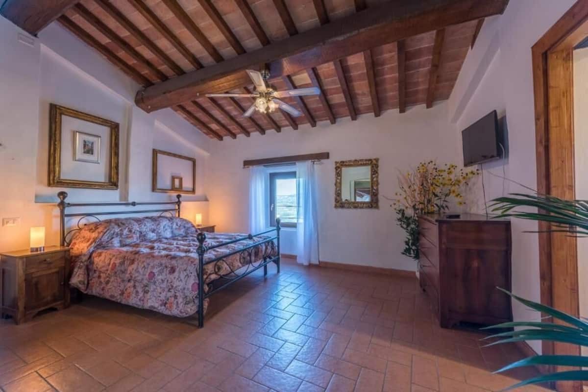 A bedroom with a wooden ceiling, a bed with a floral bedspread, a nightstand, a wooden cabinet, a television on the wall, and a window with curtains.
