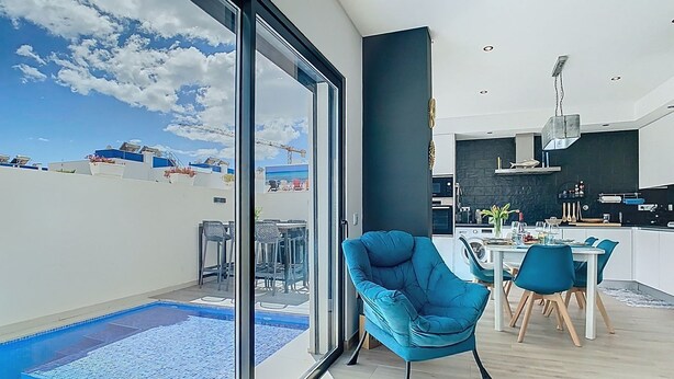 A modern living room with a large sliding glass door leading to a pool area, a dining area with blue chairs, and a kitchen with dark countertops. - Casa Launa Fuseta (Moncarapacho)