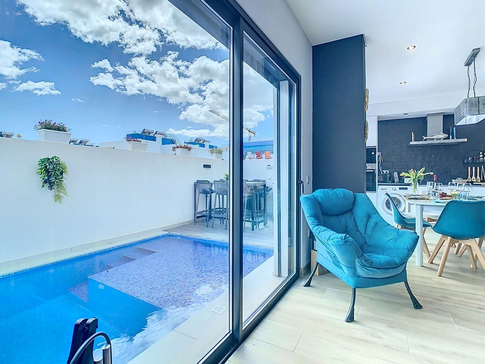 Ein modernes Wohnzimmer mit einem großen Glasfenster, das in einen Poolbereich führt, einem Essbereich und einer durch das Glas einsehbaren Küche. Im Hintergrund der Himmel und benachbarte Gebäude.