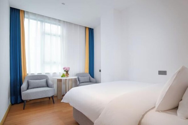 A hotel room with a large bed, two armchairs, a small round table with a vase of flowers, and a window with blue and yellow curtains.