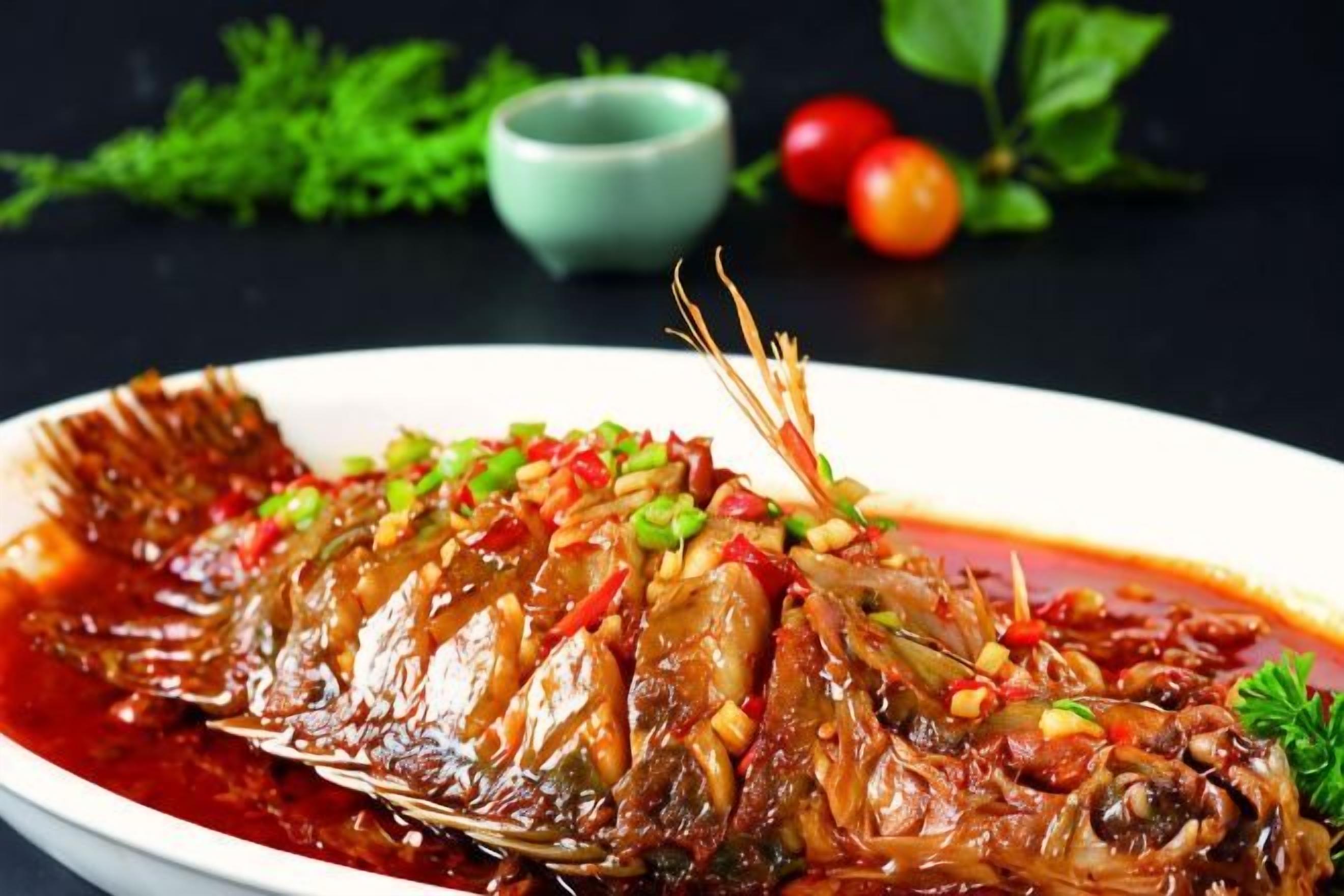 Un pescado entero cocido con salsa, adornado con cebollas verdes y pimientos rojos, sobre un plato blanco.