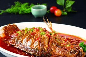 Un pescado entero cocido con salsa, adornado con cebollas verdes y pimientos rojos, sobre un plato blanco.