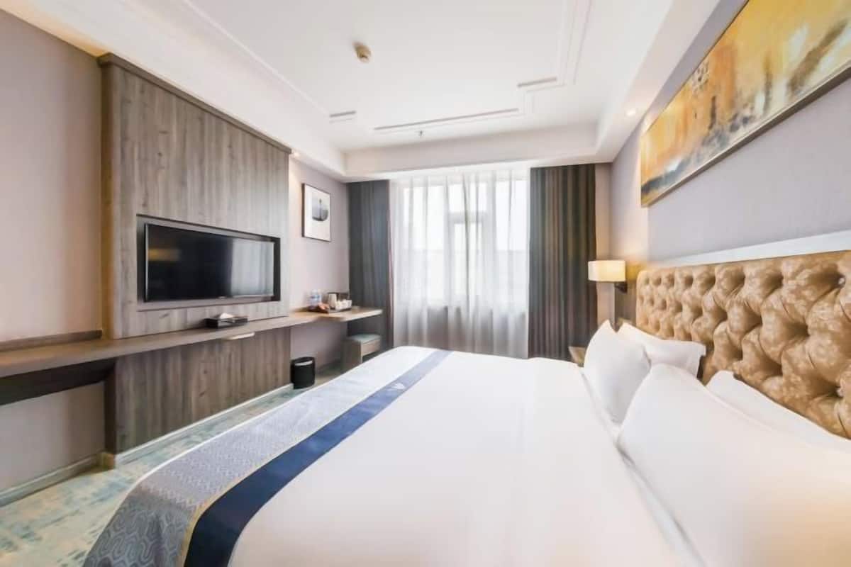 Habitación de hotel con una cama grande, un televisor de pantalla plana montado en un panel de madera, una mesita de noche y una ventana con cortinas.