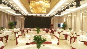 Une salle de banquet avec des tables rondes, des chaises blanches ornées de rubans rouges et un grand lustre.