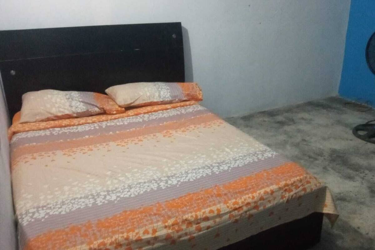 A small, single-bed room with a plain headboard, a bed with a patterned bedspread, and a standing fan against the wall.