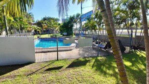 A swimming pool surrounded by a fence, with palm trees and a building in the background. - MIDTOWN CARAVAN PARK (Millbank)