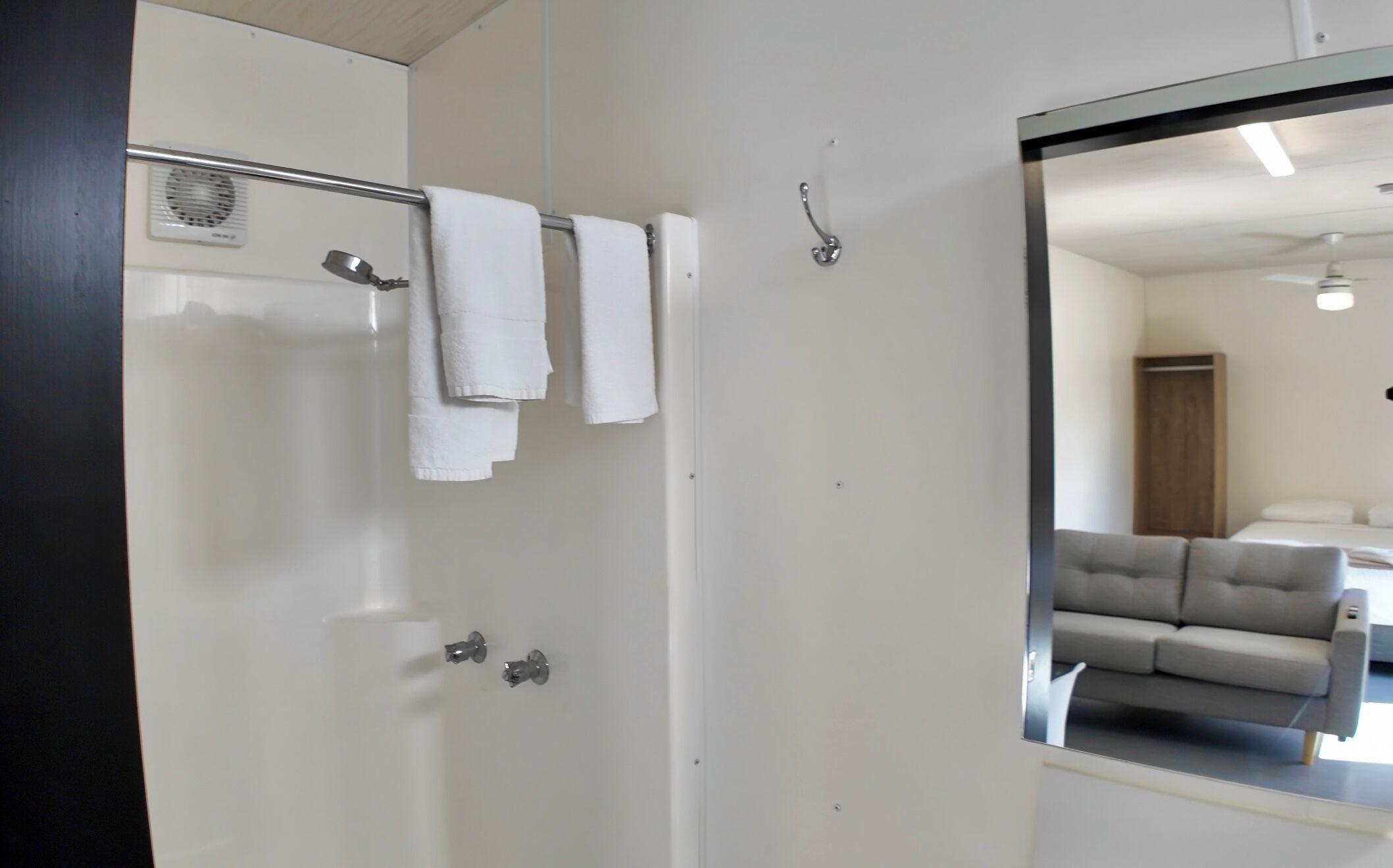 Ein Badezimmer mit Dusche, einem Handtuchhalter mit Handtüchern, einem Spiegel, der ein Wohnzimmer mit einer Couch reflektiert, und einem Deckenventilator.