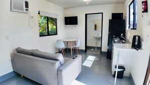 A compact living space with a grey sofa, a small round table with two chairs, a kitchenette, and a TV mounted on the wall. - MIDTOWN CARAVAN PARK (Millbank)