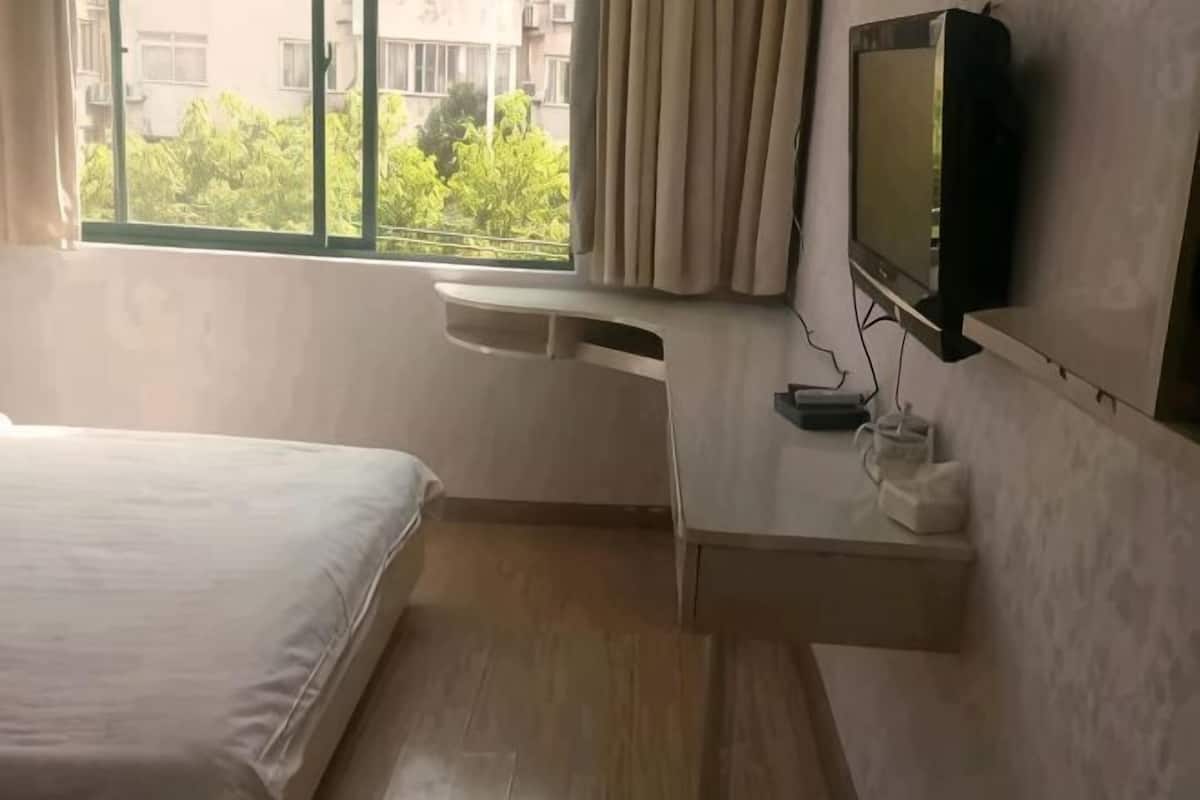 A hotel room with a bed, a TV mounted on the wall, a small table with a lamp, and a window with curtains.