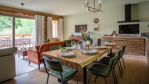 Un comedor moderno con una mesa de madera dispuesta para seis personas, rodeada de sillas tapizadas en verde. Una lámpara de araña cuelga encima, y se ve una cocina con una campana extractora negra al fondo.