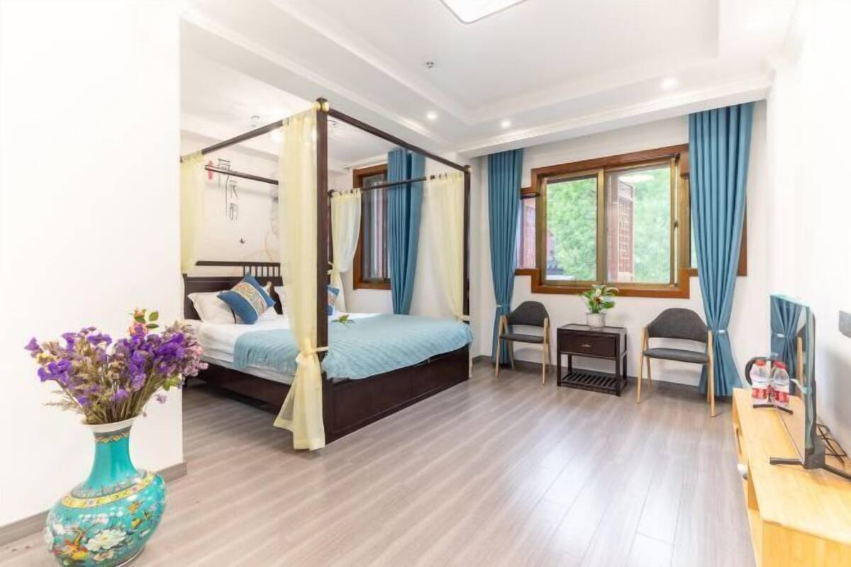 A four-poster bed with a blue bedspread, a small desk with a chair, a vase with flowers, and a window with curtains.
