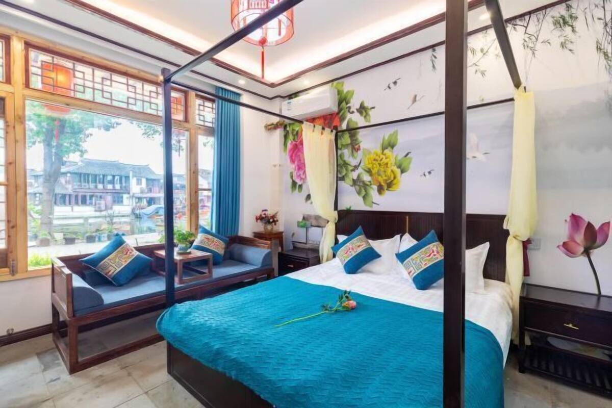 A room with a four-poster bed, a daybed, a window with a view of buildings, and a wall with floral artwork.