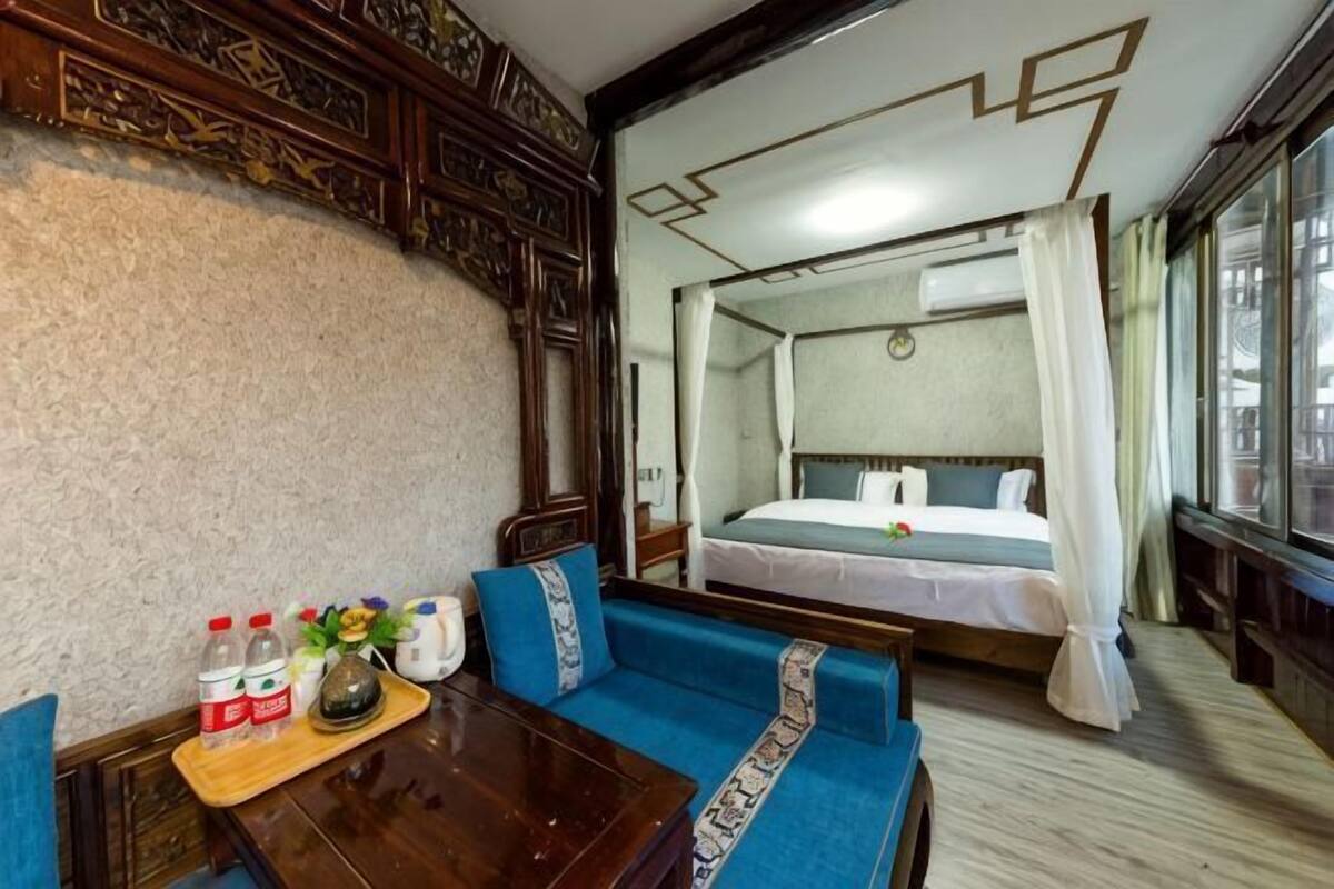 A hotel room with a bed, a sofa, a wooden table with bottles and a tray, and a large window.