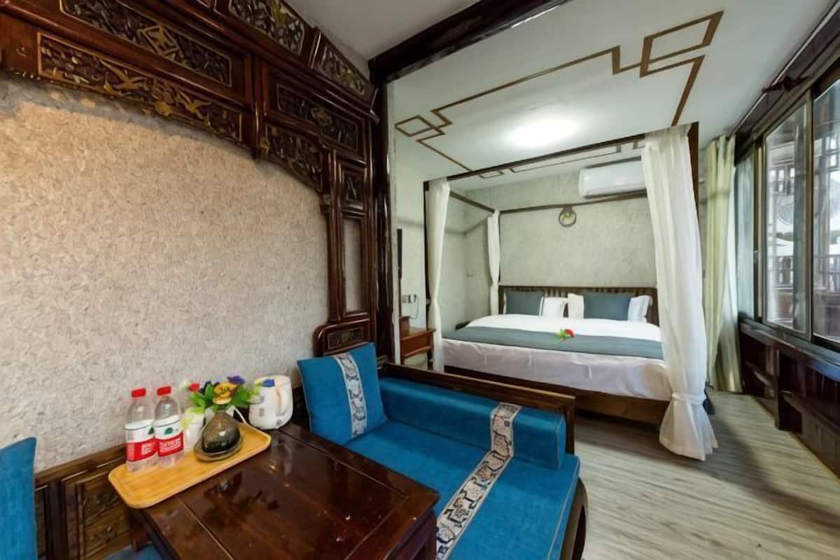 A hotel room with a bed, a sofa, a wooden table with bottles and a tray, and a large window.