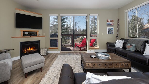 A living room with a fireplace, a flat-screen TV, a sofa, a coffee table, and a sliding glass door leading to an outdoor area with red chairs. - Winterstar Building A #6 3 bedroom views, Pet friendly common area HT (Fraser)