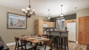 A dining area with a wooden table set for six, surrounded by chairs with cushioned seats. There is a kitchen area with wooden cabinets and a refrigerator. - Winterstar Building A #6 3 bedroom views, Pet friendly common area HT (Fraser)