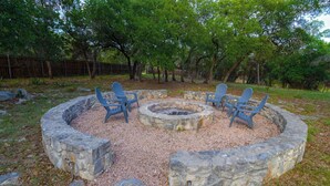 A circular stone fire pit with four Adirondack chairs around it, set in a grassy area with trees in the background. - Willie's Hill Country Hideaway - Stay for 8 With Hot Tub & Fire Pit (Wimberley)