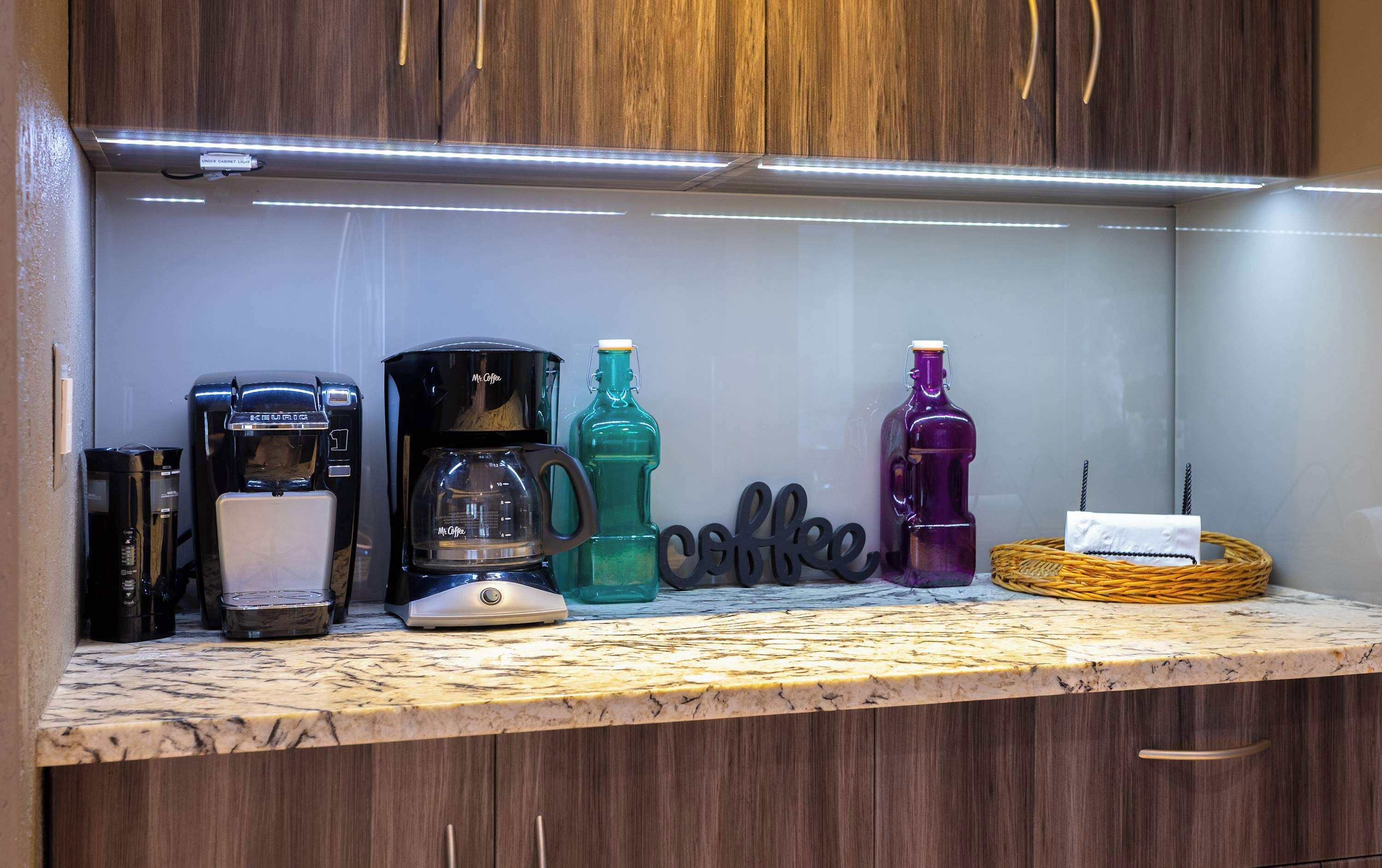 A kitchen counter with a coffee maker, a coffee pot, a green bottle, a purple bottle, and a wicker basket.