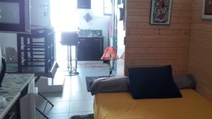 Una habitación compacta con una cama, una mesita y una silla. Se ve un área de cocina al fondo.