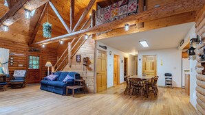 Interior de una cabaña de troncos con una escalera de madera, una sala de estar con un sofá azul, un comedor con mesa y sillas de madera, y una cocina.