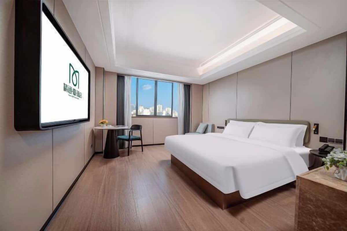 A modern hotel room with a large bed, a desk with a chair, a television mounted on the wall, and a window with a city view.