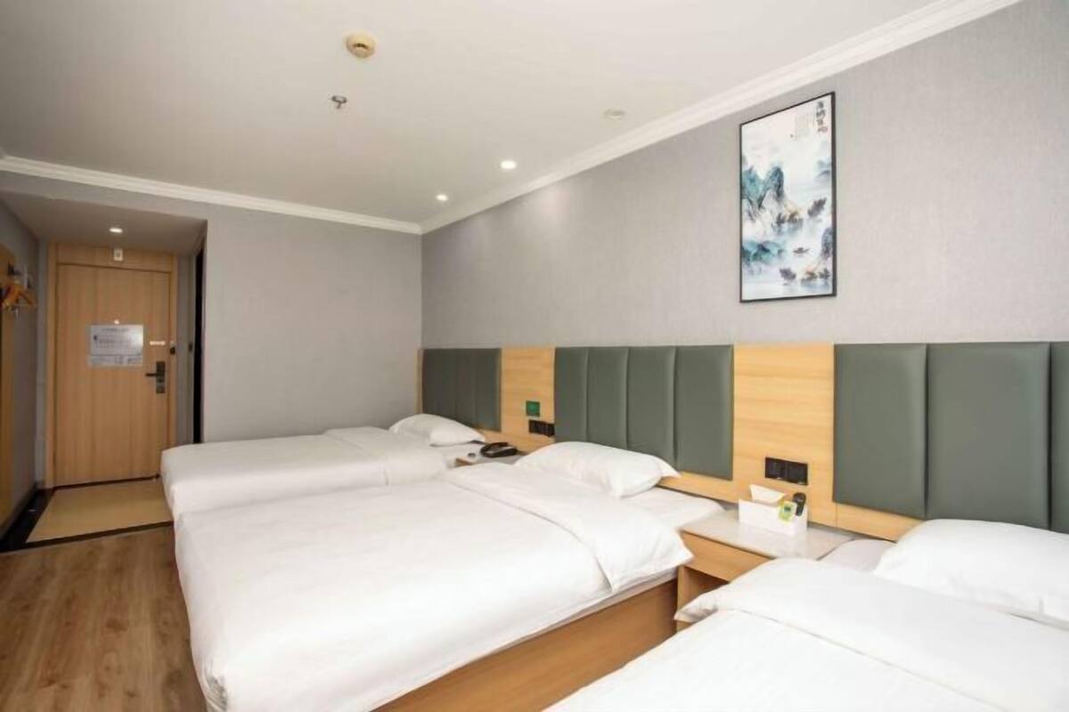 Habitación de hotel con tres camas, una puerta de madera, una obra de arte enmarcada en la pared y una mesita con un teléfono y pañuelos.