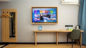 Habitación de hotel con televisor montado en la pared, un escritorio de madera con silla, perchero y equipo de aire acondicionado.