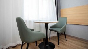 Una habitación con dos sillas verdes alrededor de una pequeña mesa redonda, un cabecero de madera y una ventana con cortinas de gasa.
