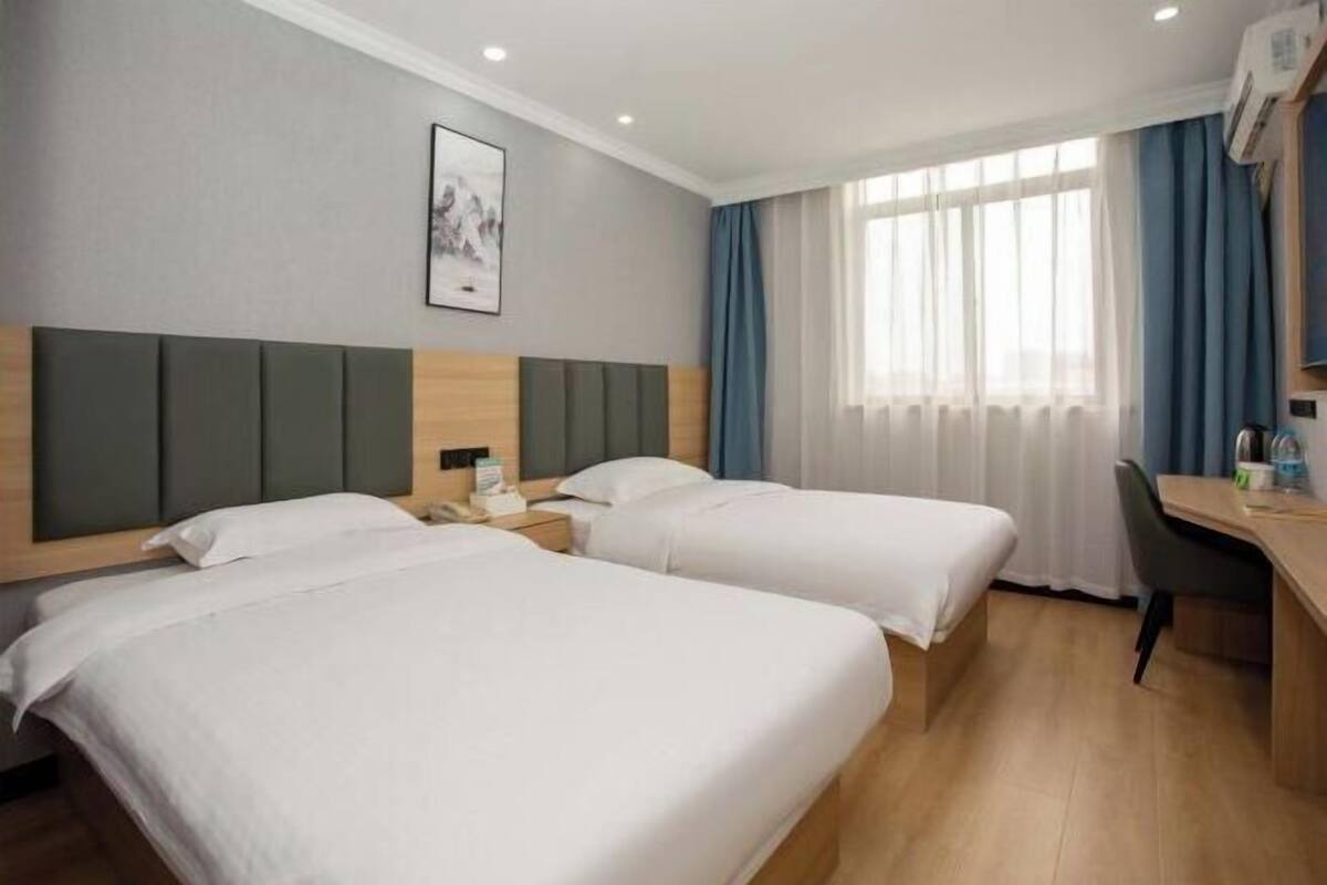 Una habitación de hotel con dos camas individuales, un escritorio, una silla, una ventana con cortinas y un cuadro enmarcado en la pared.