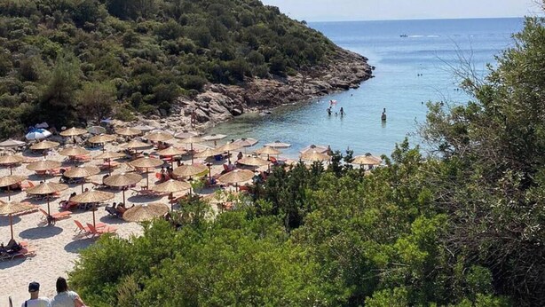 A beach with numerous thatched umbrellas and sun loungers, surrounded by dense greenery and rocky coastline.