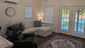 A living room with a sectional sofa, a dining table, and a patterned rug. There are two windows with blinds, a clock on the wall, and a door leading outside. - The Hawthorne Place (Buena Vista)