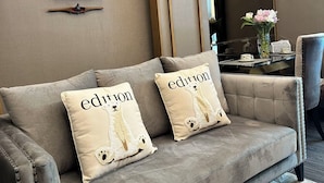 Una sala de estar moderna con un sofá gris, dos cojines con el texto 'edition', una obra de arte enmarcada y un jarrón con flores sobre una mesita auxiliar.