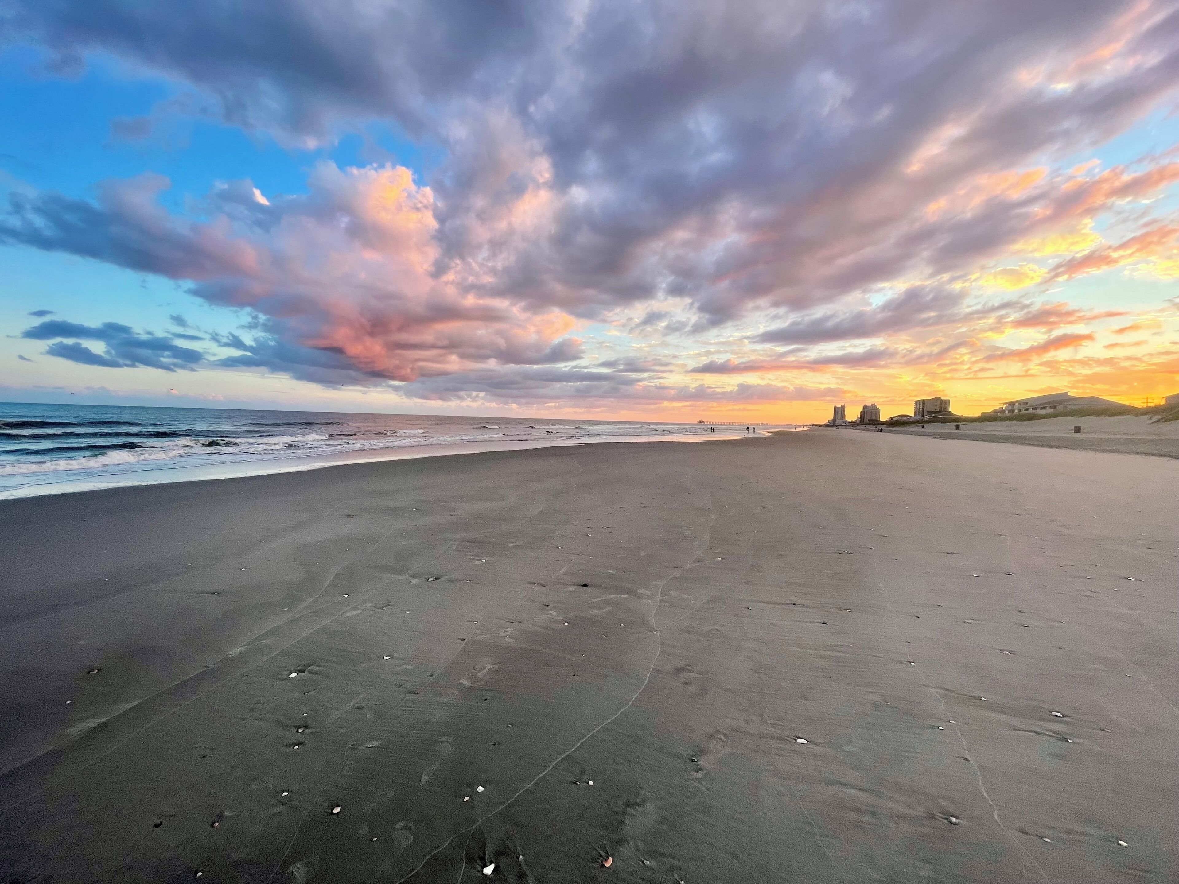 A beach with a wide expanse of sand, a few scattered footprints, and a distant view of buildings and hills under a colorful sunset sky.