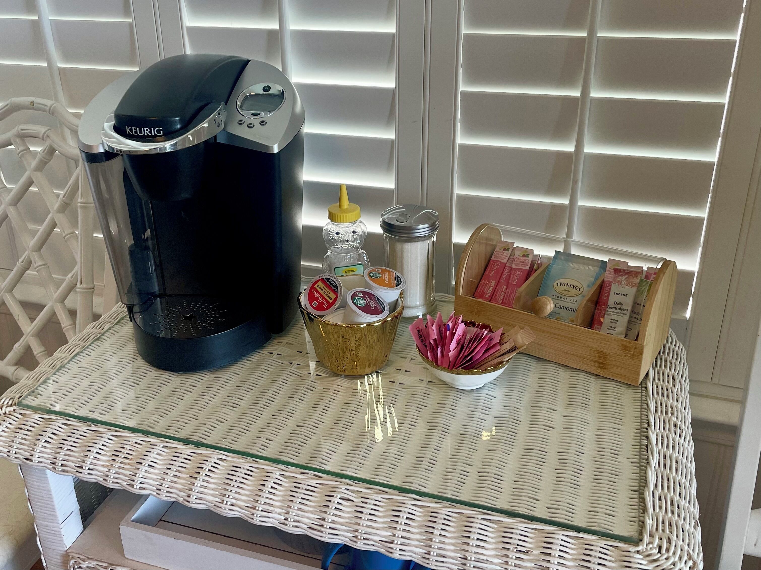 A coffee maker, a honey dipper, a small bowl of honey, a honey dipper, and a wooden box with tea bags on a wicker table.