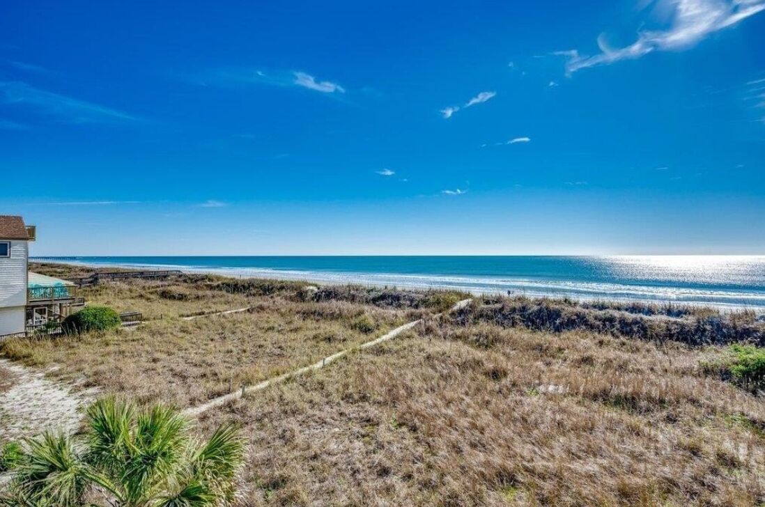 Une propriété située sur la côte, avec un sentier sablonneux menant à une plage, une maison dotée d’une terrasse offrant une vue sur l’océan et un ciel d’un bleu limpide.