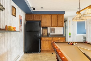 A kitchen with wooden cabinets, a black refrigerator, a pool table, and a ceiling light fixture.