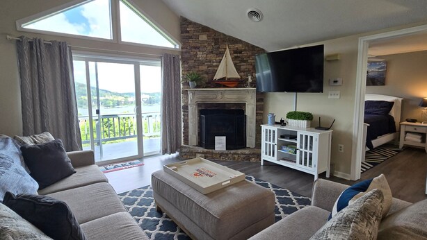 A spacious living room with a fireplace, a large TV, and a view of a lake through the sliding glass doors.