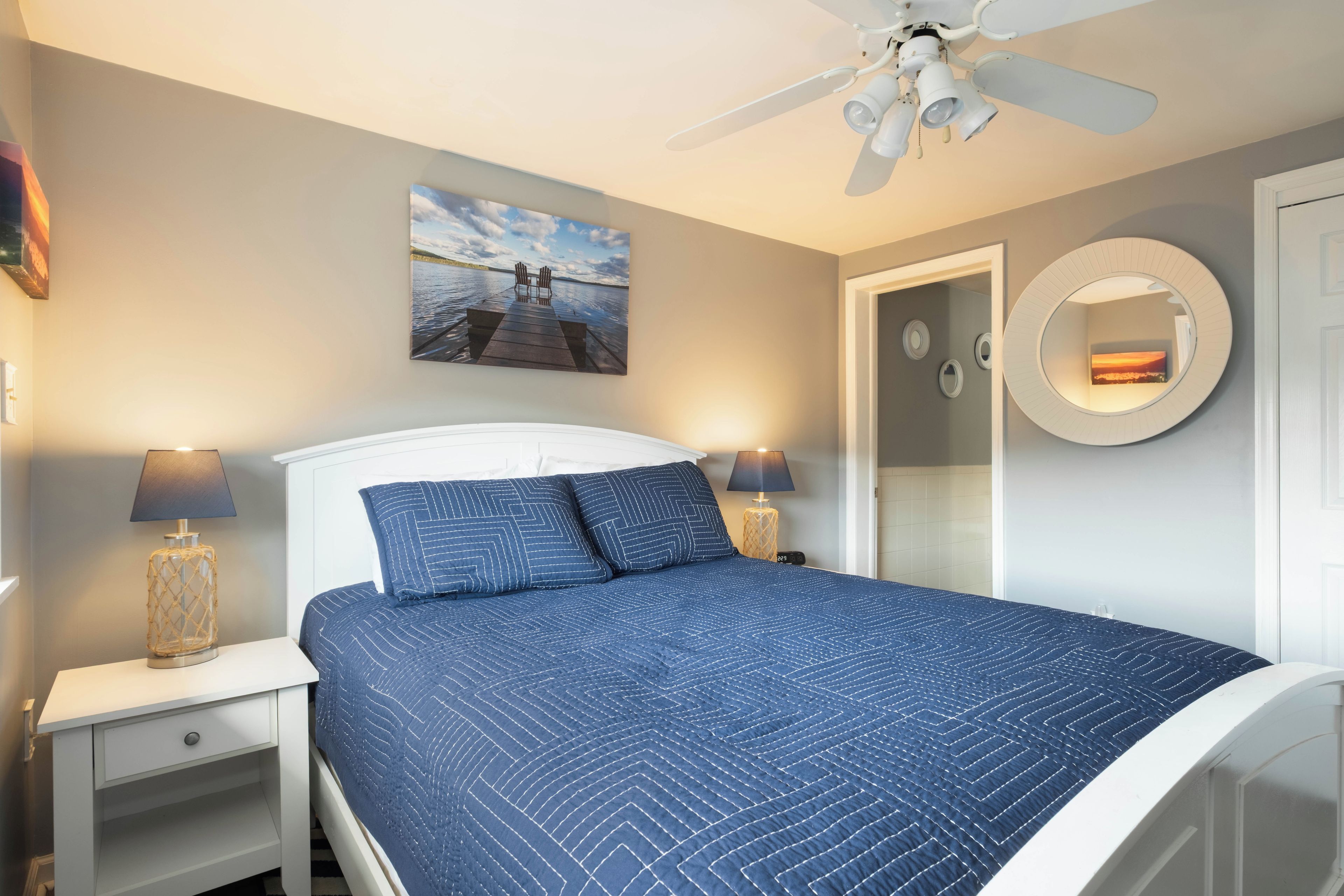 Una camera da letto con un letto, un ventilatore a soffitto, un quadro, uno specchio rotondo e un comodino con una lampada.