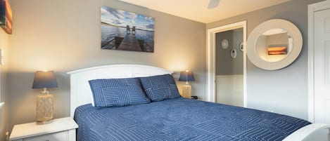 A bedroom with a bed, a ceiling fan, a painting, a round mirror, and a nightstand with a lamp.