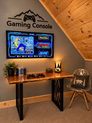 A gaming console area with a TV displaying a game menu, a wooden table with gaming accessories, and a chair.