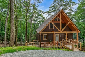 A cabin with a steeply pitched roof, a front porch with a railing, and a gravel driveway.