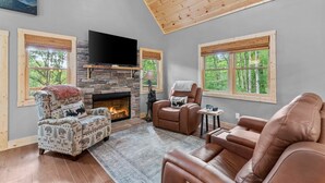 A cozy living room with a stone fireplace, leather sofas, and large windows offering a view of trees. - Bear Necessities - Hot tub, Pet Friendly, Games, Fire table, Beech Mtn (Beech Mountain)