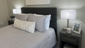 Een net opgemaakt bed met een grijze deken en kussens, een donker hoofdbord, een nachtkastje met een lamp en een fotolijstje, en een aan de muur bevestigd abstract kunstwerk.