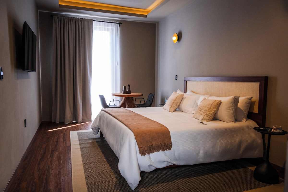 Habitación de hotel con una cama grande, un televisor montado en la pared, una mesita con sillas y una ventana con cortinas.