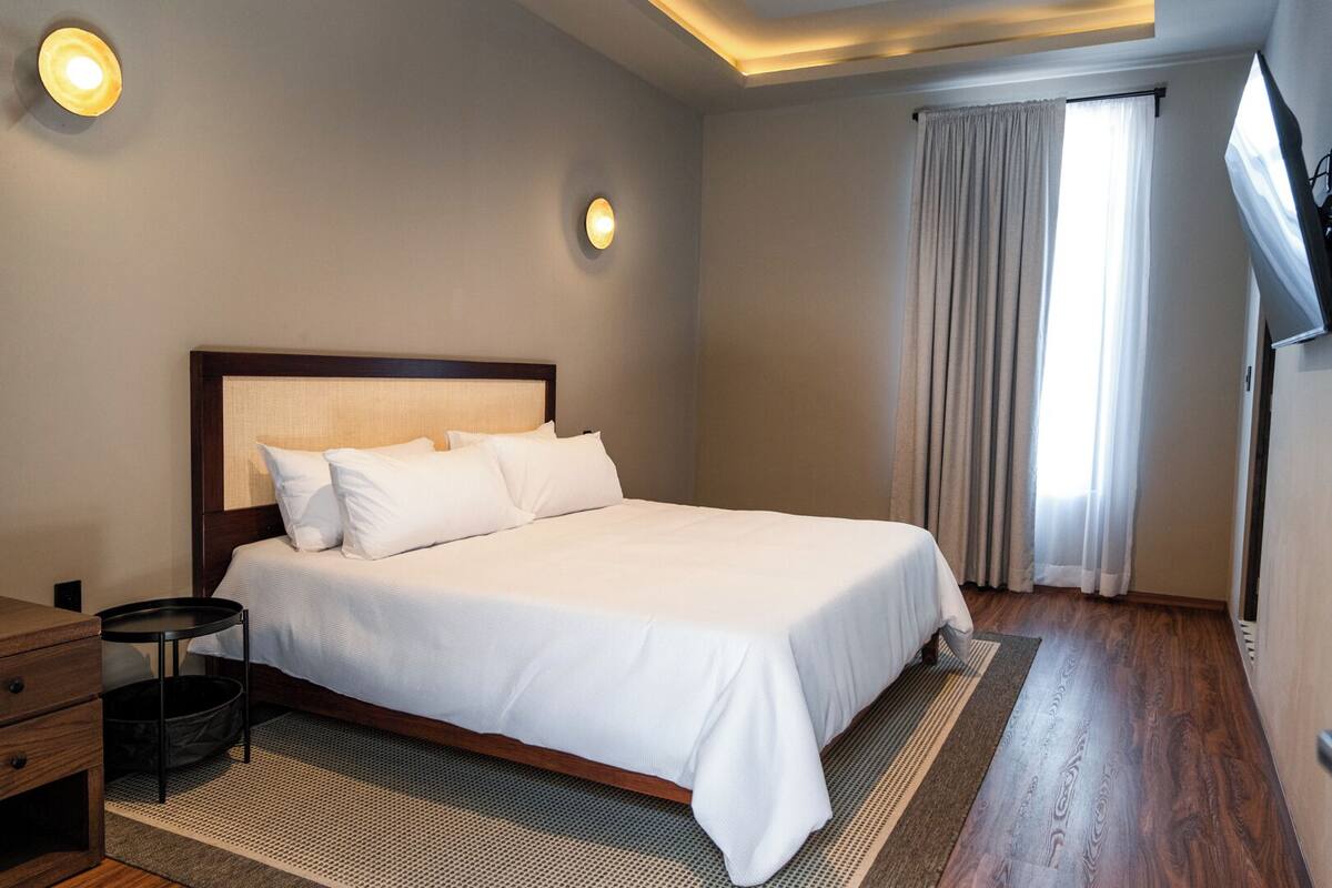 Una cama cuidadosamente tendida en una habitación de hotel, con cabecera de madera, una mesita de noche pequeña y un televisor en la pared.