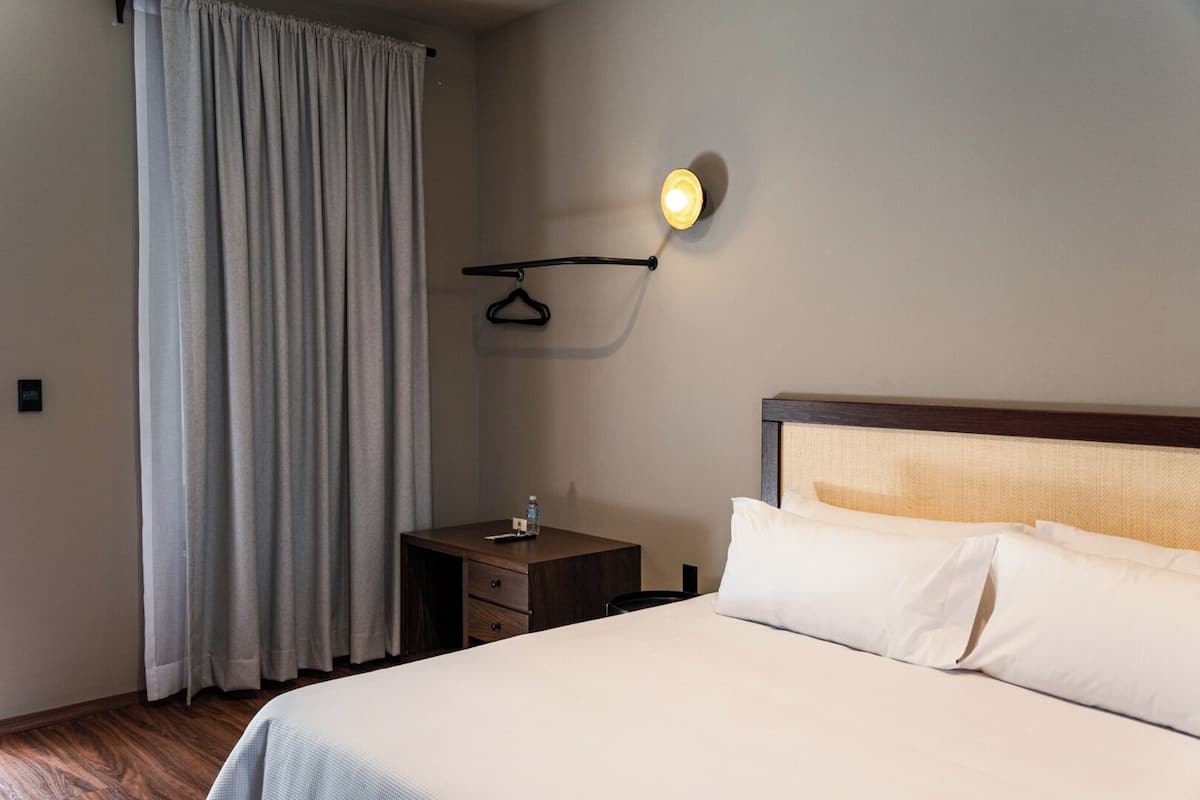 Una habitación de hotel con cama, mesita de noche, lámpara colgante y ventana con cortinas.