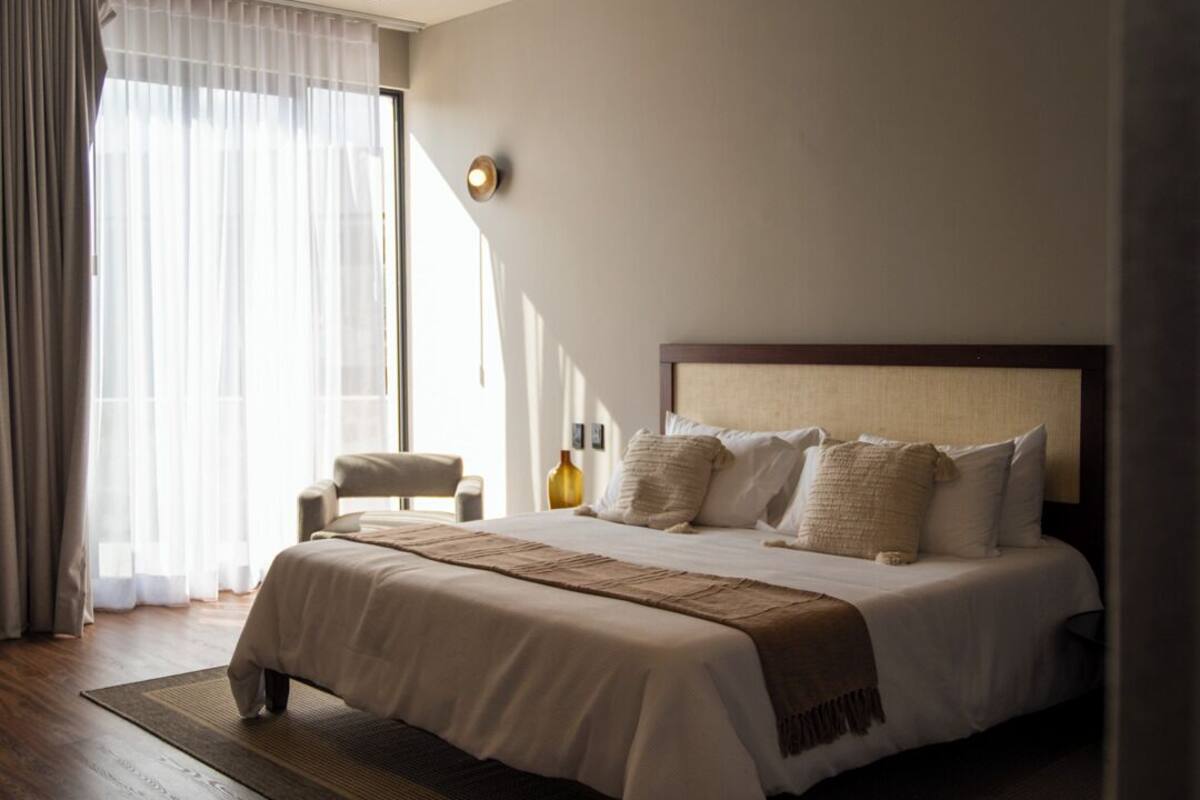 Una habitación de hotel moderna con una cama grande, piso de madera y un ventanal con cortinas translúcidas.